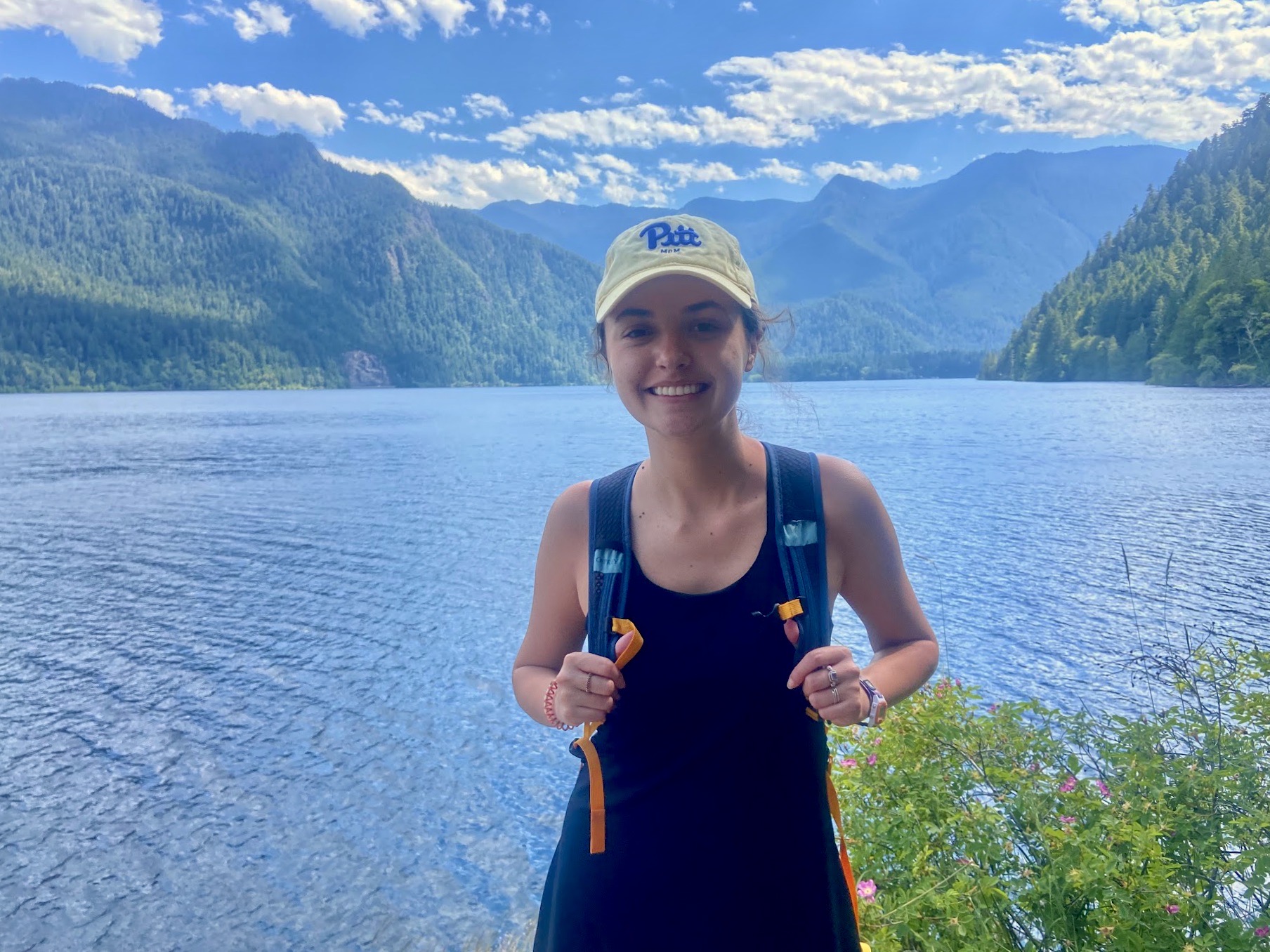 Me standing in front of a teal lake in Washington wearing a 
        yellow 'Pitt' baseball cap and a backpack.